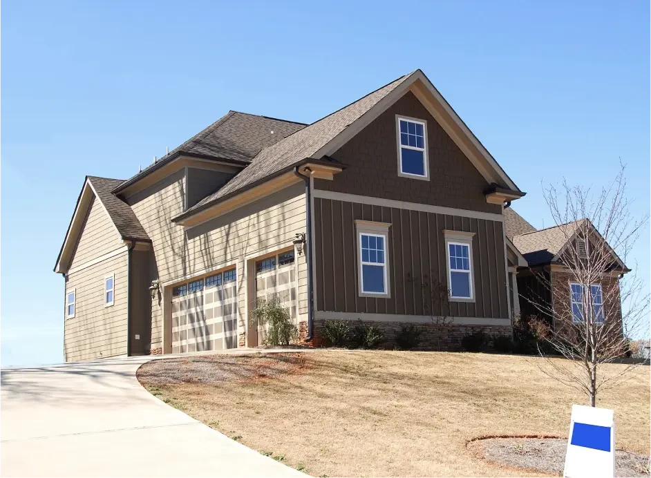 Dom na wzieniesieniu w brązowych odcieniach. Ściany wykończone panelami drewnianymi, dach – gontami. Betonowy podjazd oraz równo przystrzyżony trawnik pokazują, że posiadłość jest schludna i zadbana