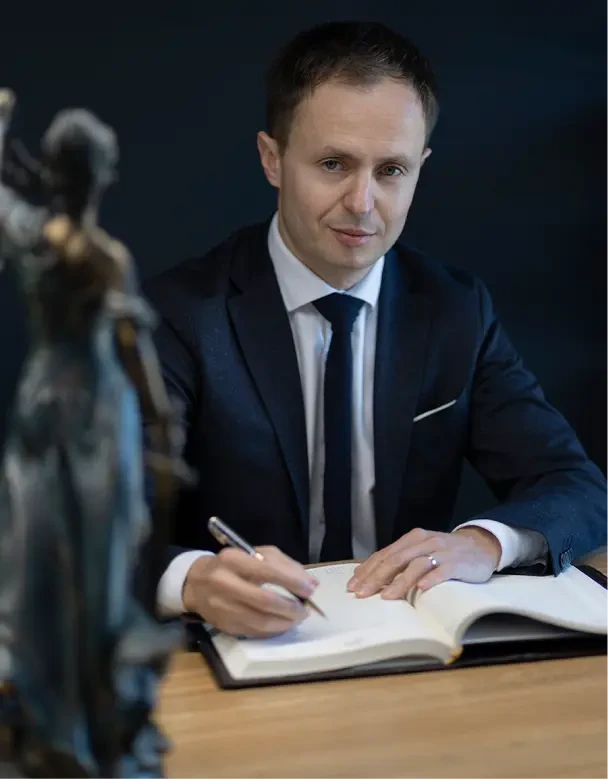 Piotr Nowakowski w czarnym garniturze i białej koszuli. Lekko uśmiechnięty mężczyzna, elegancko ubrany patrzy w obiektyw. Na pierwszym planie rozmazana figurka Temidy – greckiej bogini symbolizującej prawo i wieczny porządek.