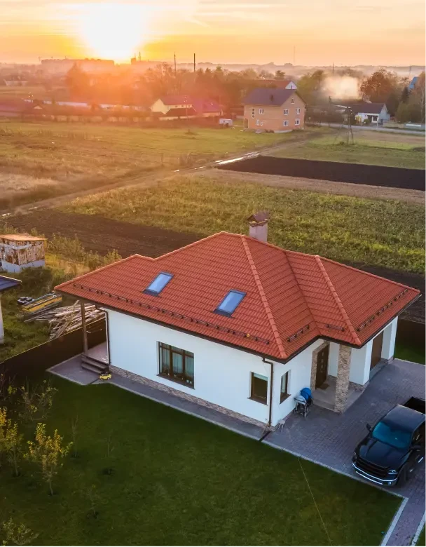 Widok z drona na nieruchmość – dom z działką i zaparkowanym samochodem na podjeździe. W tle widać zielone tereny i inne domy, działki i pola oraz zachodzące słońce